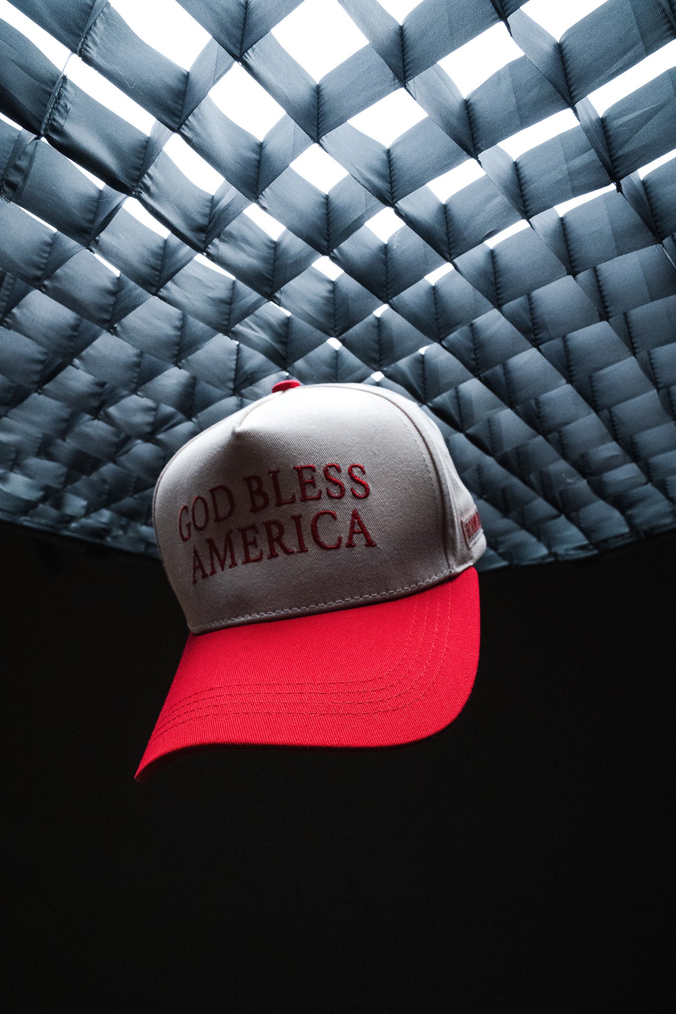 A red and white Born United Private Label “God Bless America Snapback,” featuring bold front lettering, hangs beneath a grid-patterned light.