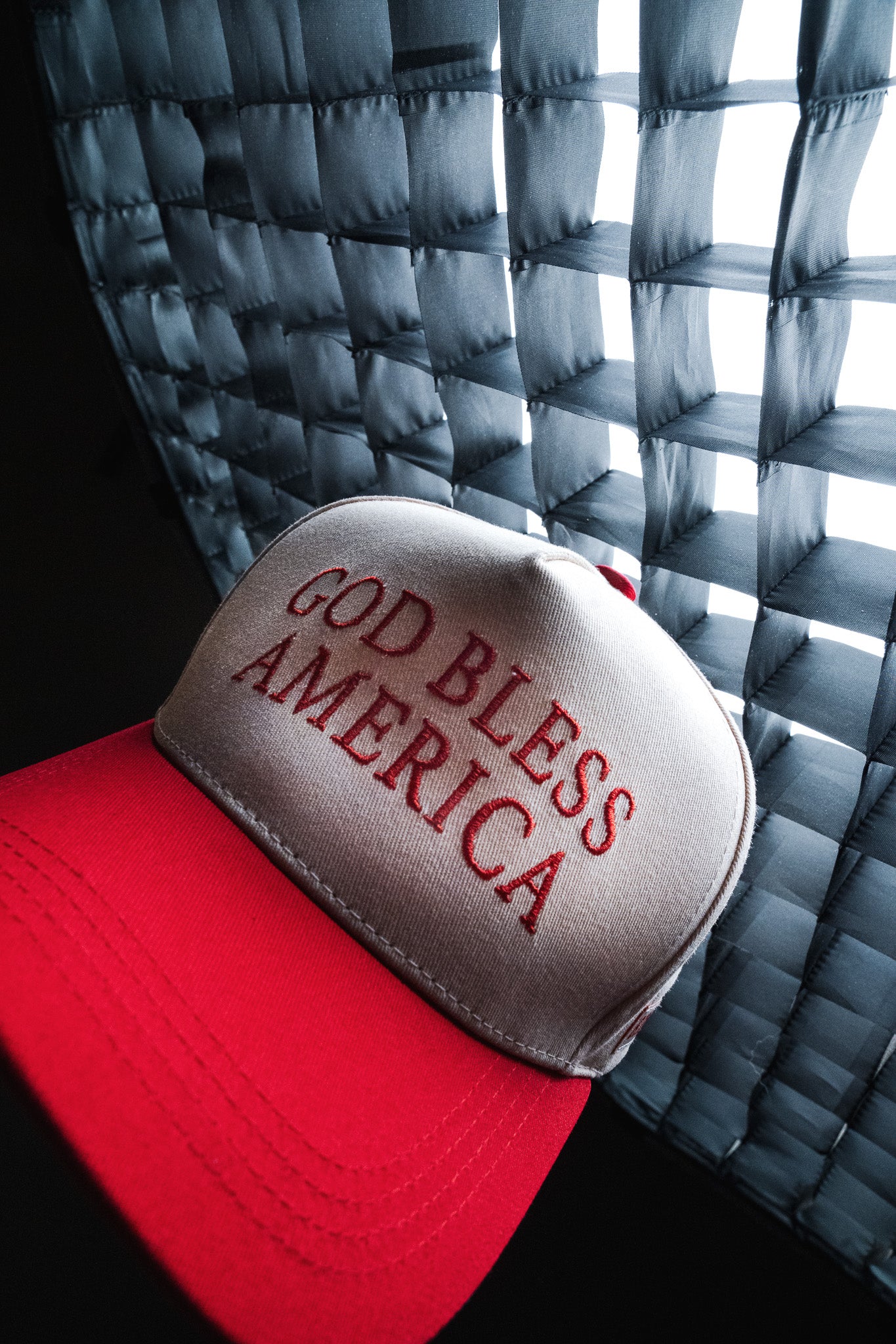 Born United Private Label’s God Bless America Snapback, a red and white embroidered hat featuring “GOD BLESS AMERICA” in bold red lettering, is pictured near a window with sunlight filtering through blinds.