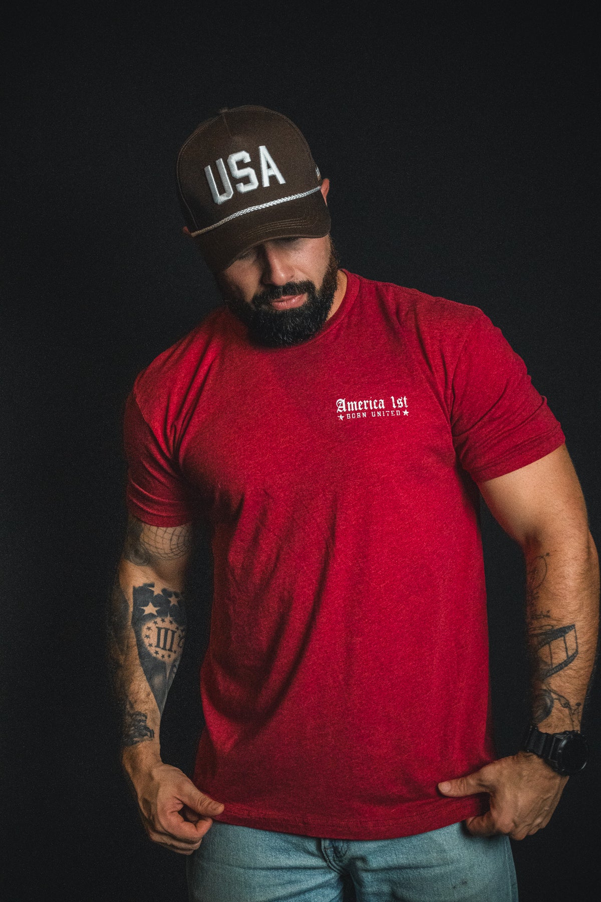 A man with a beard, wearing a red America 1st t-shirt, jeans, and a Born United Private Label USA Brown Snapback Hat stands against a black background.