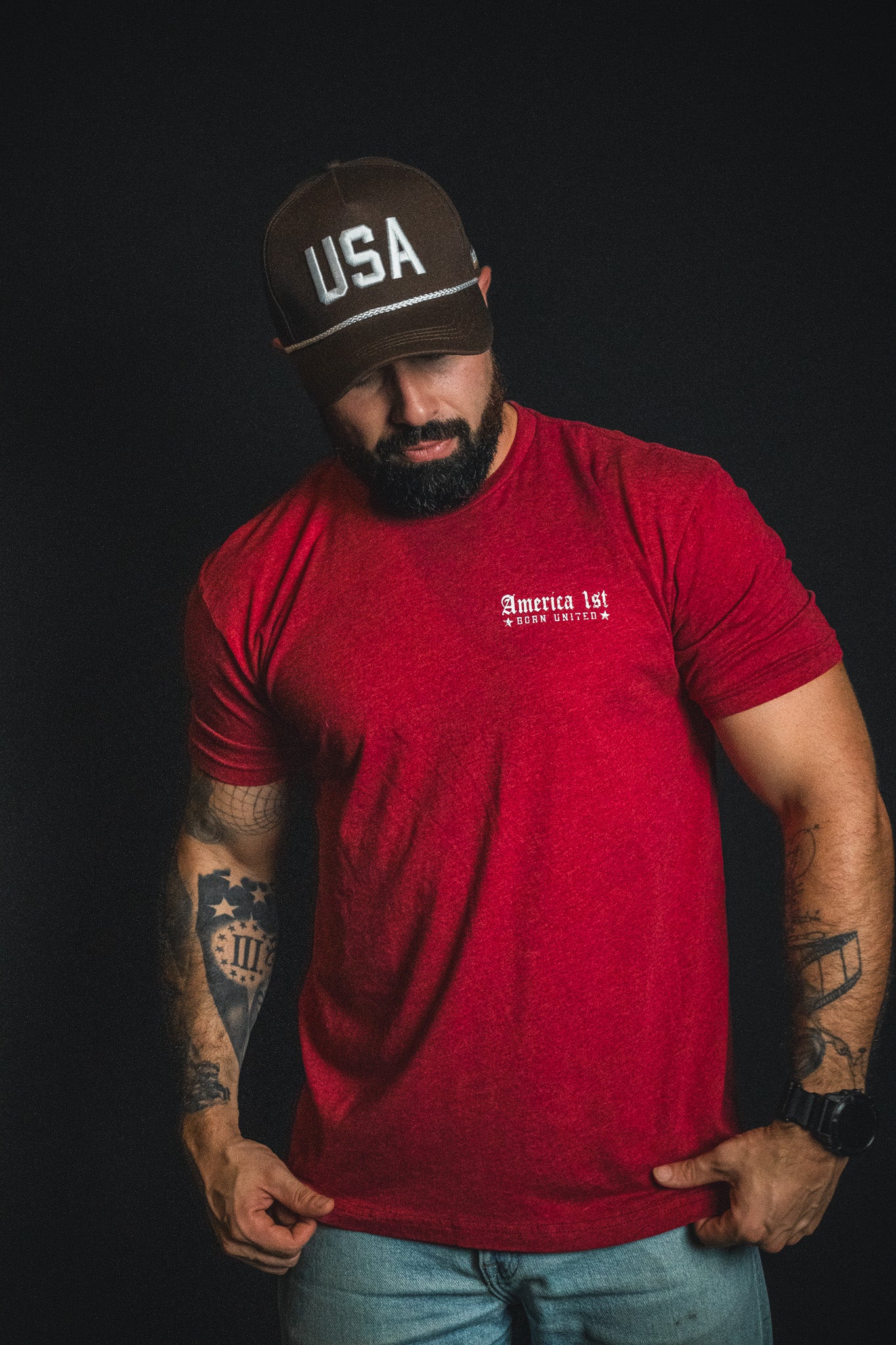 A man with a beard, wearing a red America 1st t-shirt, jeans, and a Born United Private Label USA Brown Snapback Hat stands against a black background.