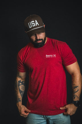 A man with a beard, wearing a red America 1st t-shirt, jeans, and a Born United Private Label USA Brown Snapback Hat stands against a black background.