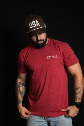 A man wearing the Born United Private Label USA Brown Snapback Hat and a red America 1st t-shirt stands against a black background, looking downward with one hand holding his shirt.