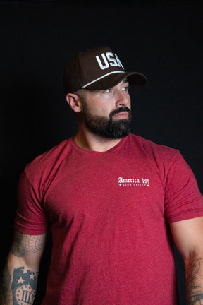 A man with a beard, wearing a Born United Private Label USA Brown Snapback Hat and a red America 1st Born Rotten t-shirt, stands against a black background.