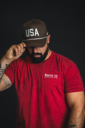A man in a red America 1st t-shirt and a Born United Private Label USA Brown Snapback Hat tilts his head down, touching the brim of his hat, against a plain dark background.