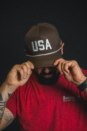 A person in a red America 1st shirt adjusts a Born United Private Label USA Brown Snapback Hat, head bowed, against a dark background.