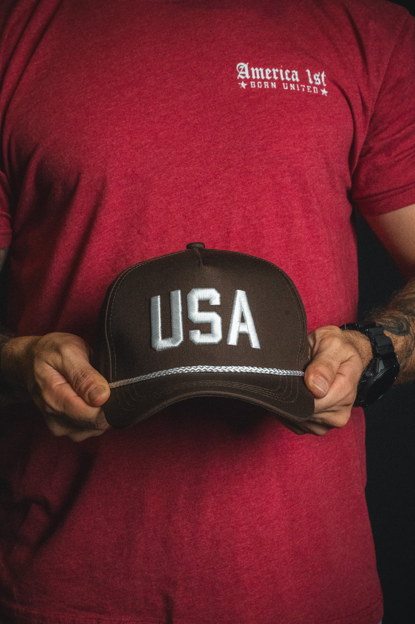A person in a red America 1st Born United shirt holds a brown USA snapback hat by Born United Private Label, featuring large white letters across the front.