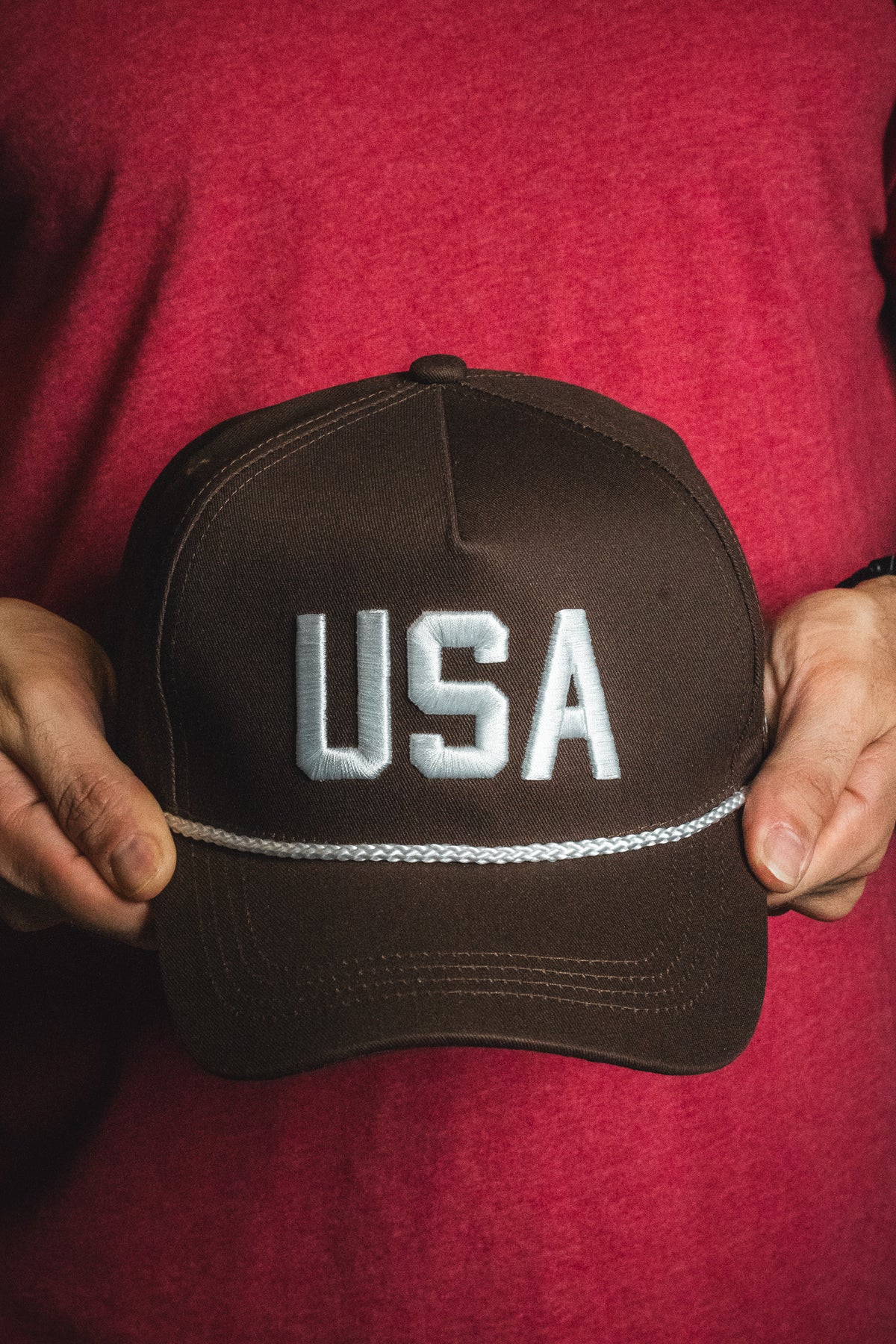 A person holding the Born United Private Label USA Brown Snapback Hat, featuring large white embroidered letters on the front, stands against a red shirt background.