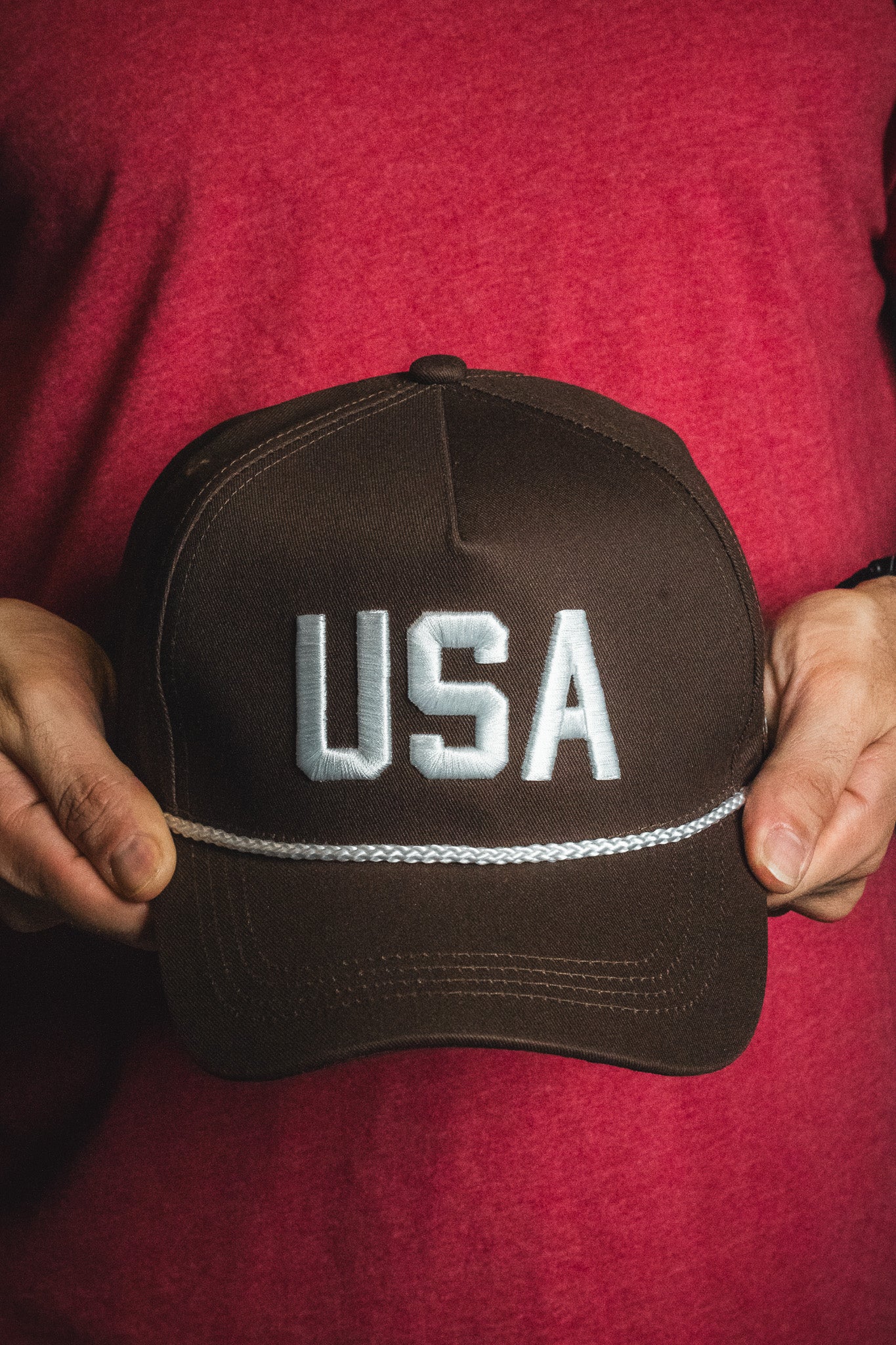 A person holding the Born United Private Label USA Brown Snapback Hat, featuring large white embroidered letters on the front, stands against a red shirt background.