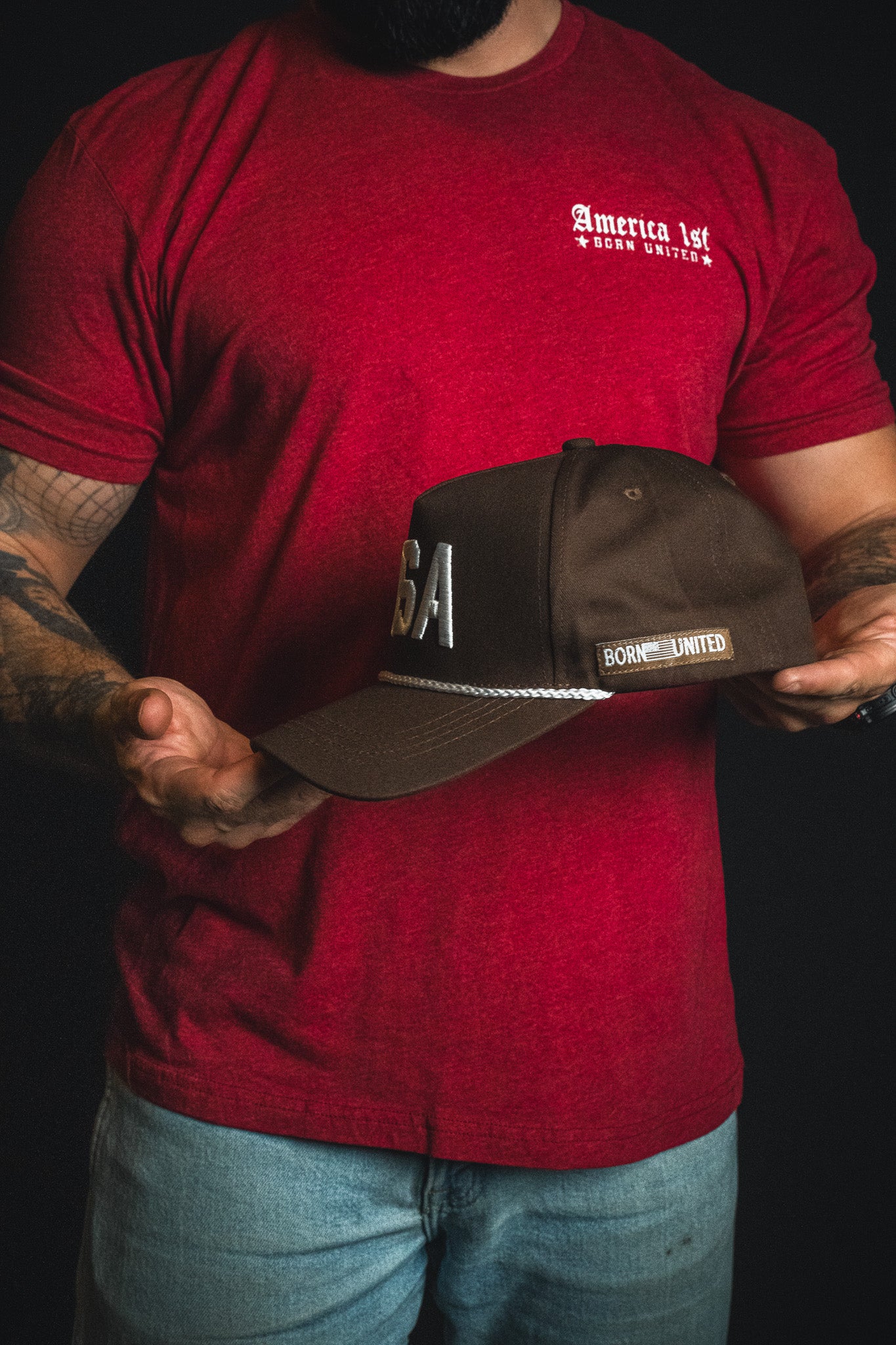 A man in a red America 1st t-shirt and jeans holds the USA Brown Snapback Hat by Born United Private Label, featuring dark brown embroidery and “USA and Born United” detailing, styled as a classic USA snapback, against a black background.