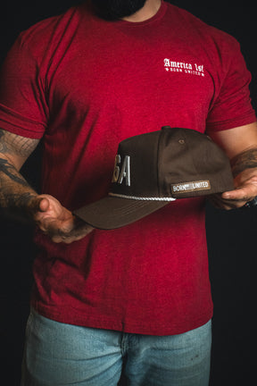 A man in a red America 1st t-shirt and jeans holds the USA Brown Snapback Hat by Born United Private Label, featuring dark brown embroidery and “USA and Born United” detailing, styled as a classic USA snapback, against a black background.