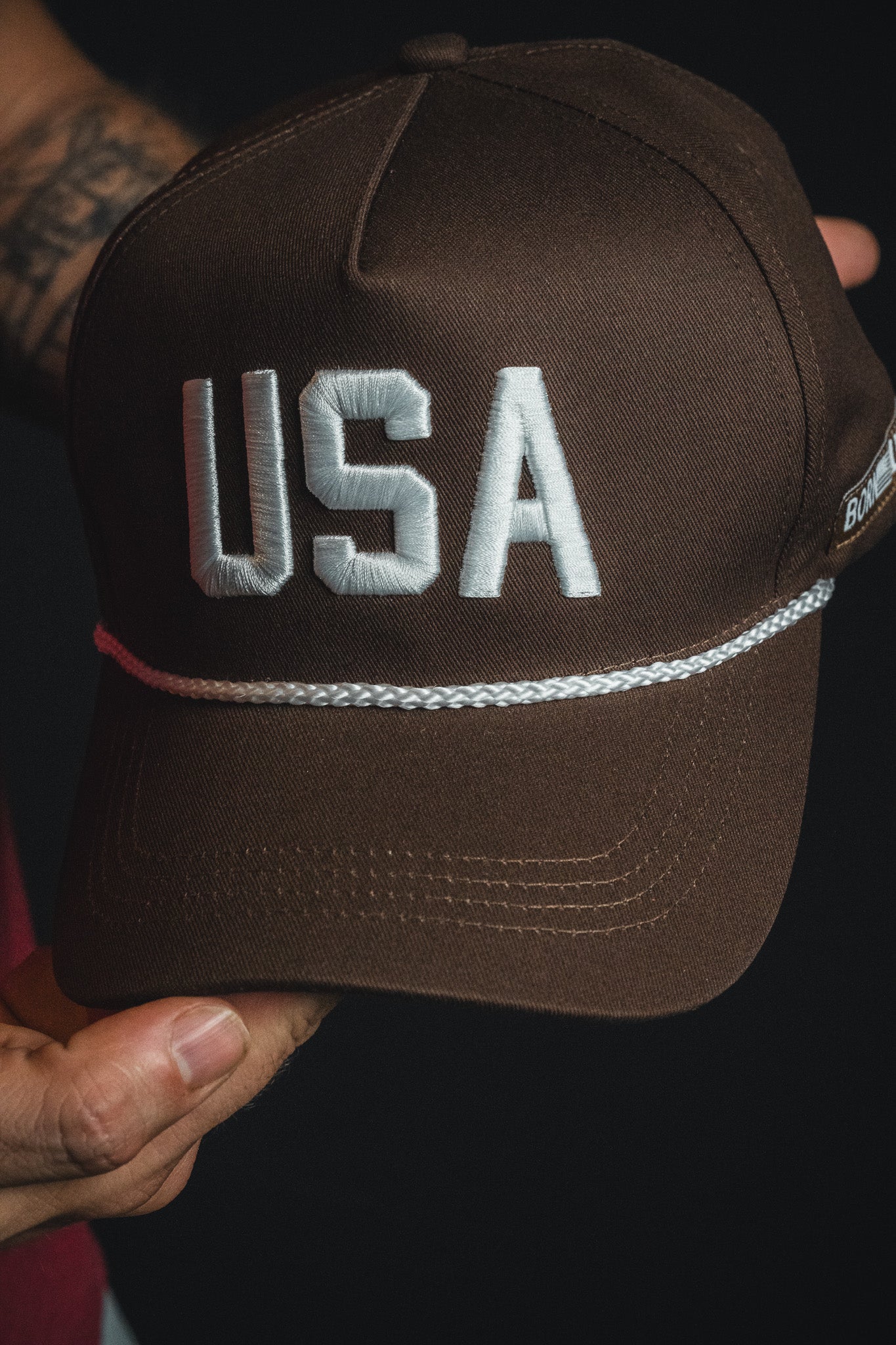 A person holds the Born United Private Label USA Brown Snapback Hat, featuring “USA” in large white letters on the front and a white cord above the brim.