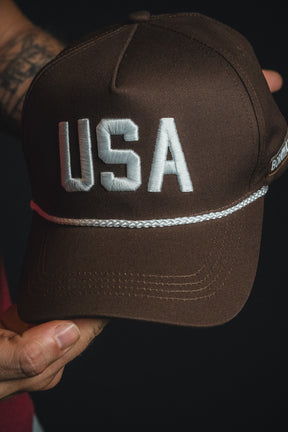 A person holds the Born United Private Label USA Brown Snapback Hat, featuring “USA” in large white letters on the front and a white cord above the brim.