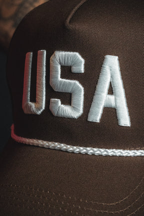 A close-up of the Born United Private Label USA Brown Snapback Hat, featuring “USA” embroidered in white on the front and a white braided band above the brim.