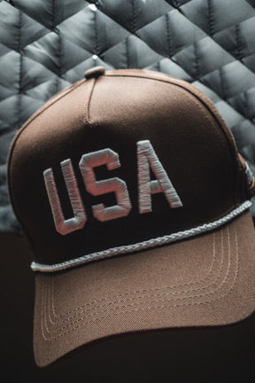 The USA Brown Snapback Hat by Born United Private Label features white “USA” embroidery on the front and is displayed against a quilted gray background.