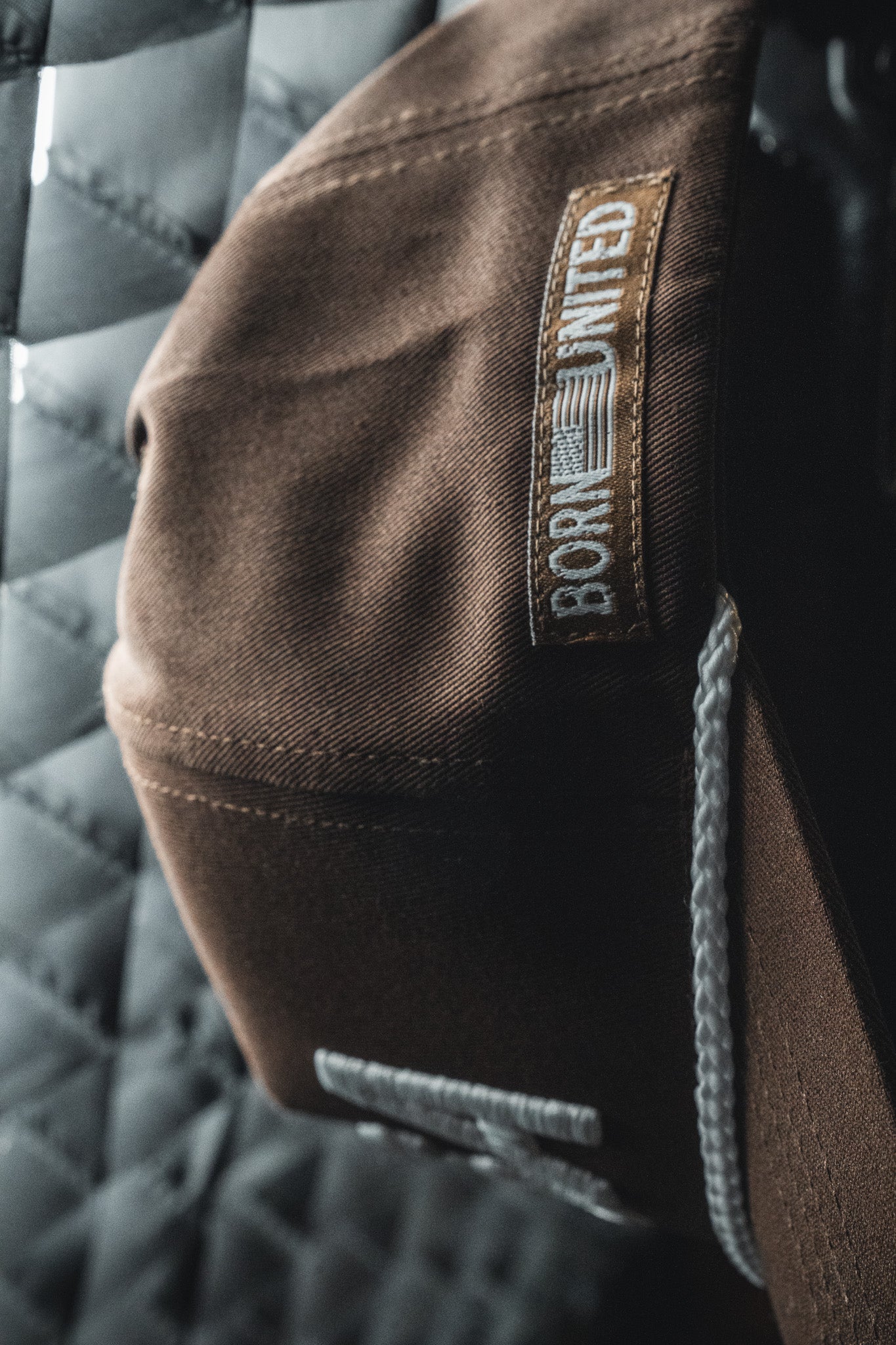 Close-up of the USA Brown Snapback Hat by Born United Private Label, featuring an embroidered Born United patch and an American flag, hanging in front of a quilted metallic surface.