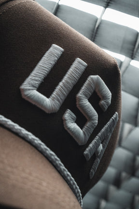 Close-up of the Born United Private Label USA Brown Snapback Hat, featuring large white embroidered “USA” lettering and a braided trim along the brim.
