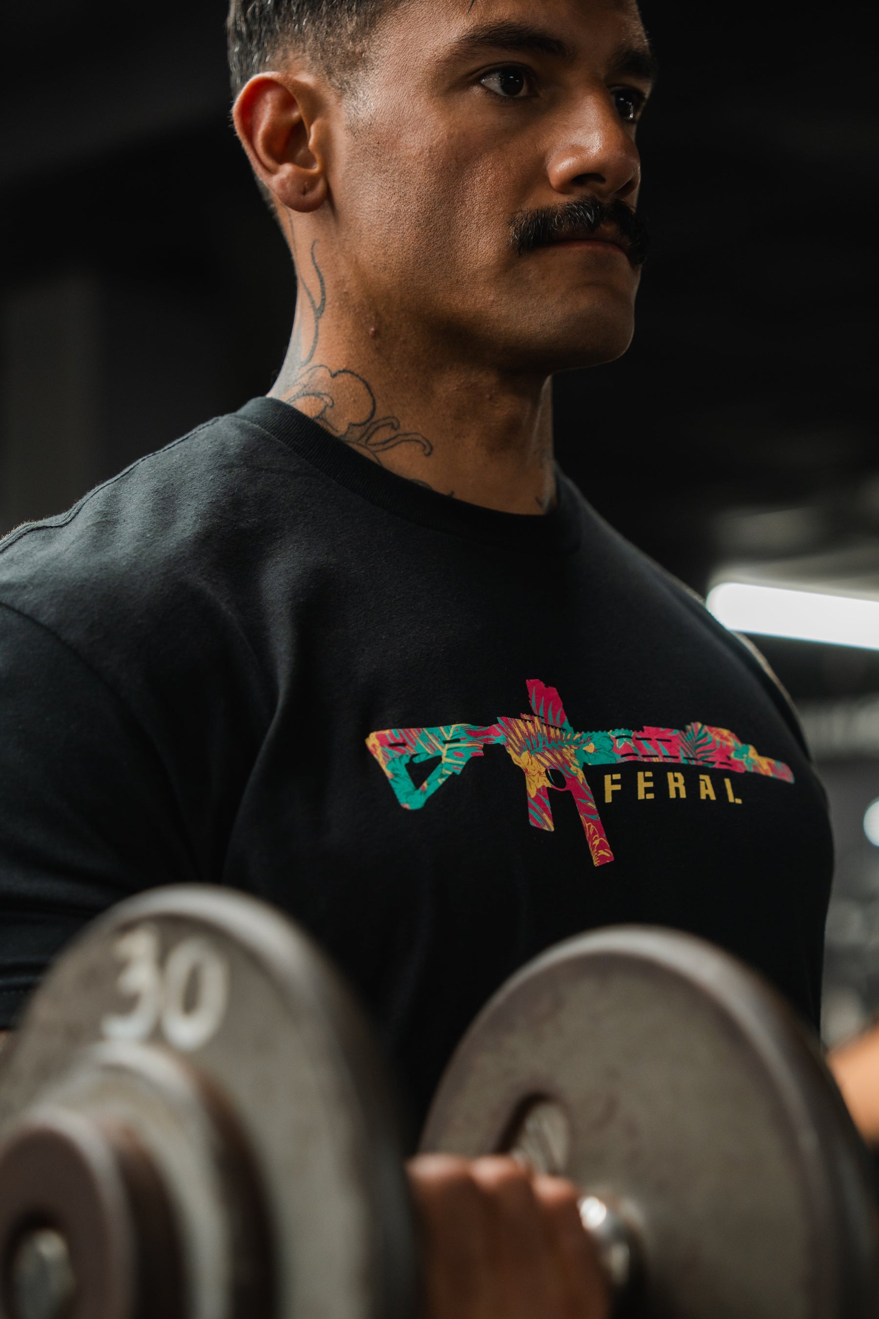 A man lifts 30-pound dumbbells, wearing the Feral - Tee by Tango Charlie Apparel. The black shirt features a colorful rifle graphic and "FERAL" text. A blurred background highlights his authentic presence and the tee’s bold design.