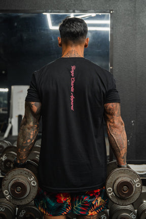 A muscular, tattooed man in a Tango Charlie Apparel "Feral" Tee and floral shorts stands confidently in the gym with heavy dumbbells—mirrors and weights reflecting his radical authenticity.
