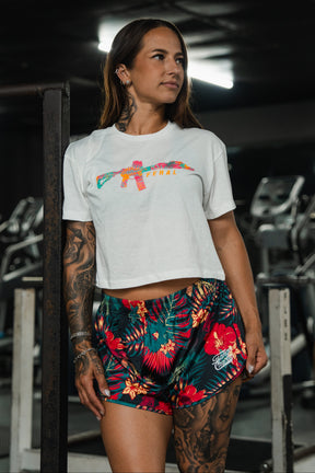 A tattooed woman stands in a gym, wearing the Tango Charlie Apparel Feral - Women's White Crop with a colorful graphic, paired with dark floral shorts. She looks to the side, gym equipment visible behind her.