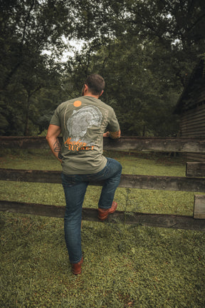 A man in Mucker Bucket’s green Ghost Gobbler Premium Tee and jeans climbs a wooden fence in a grassy, wooded area by a rustic building.
