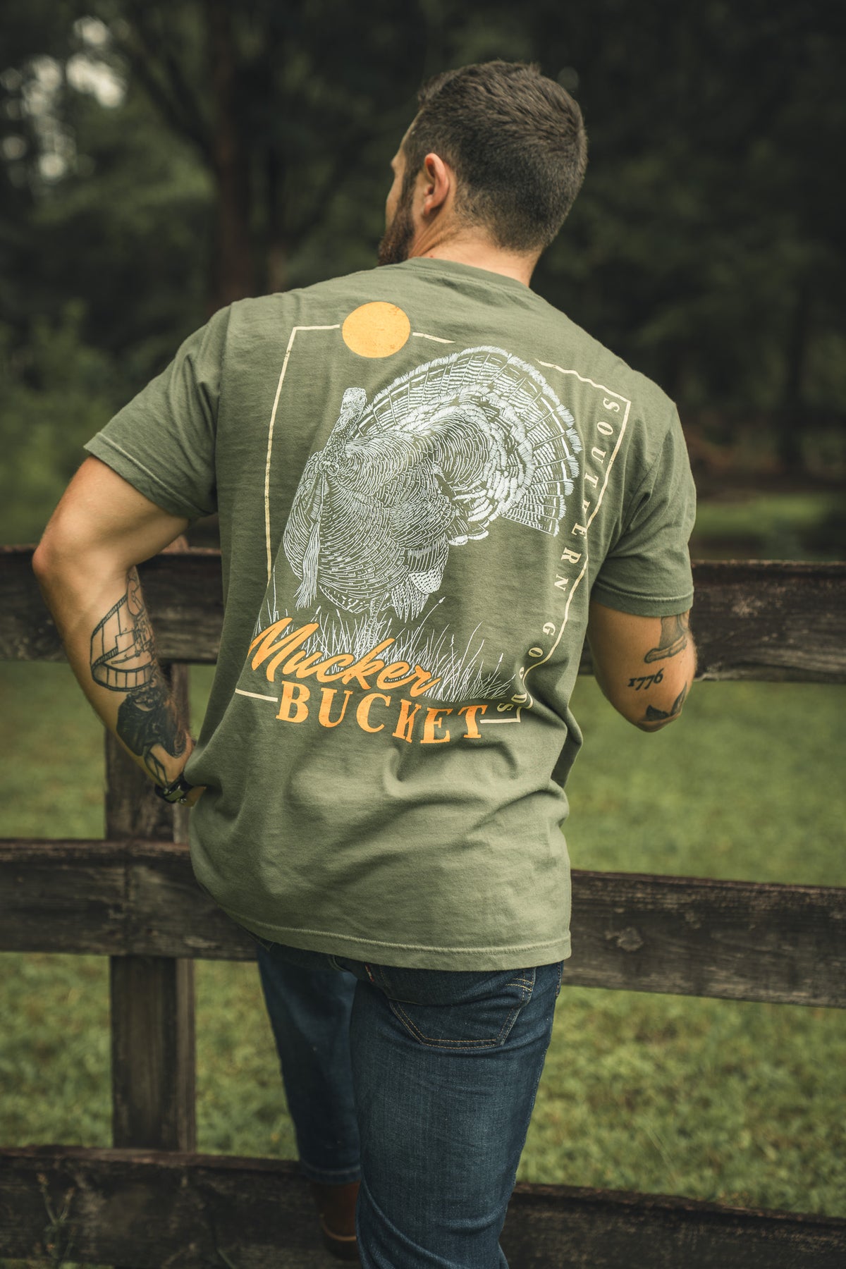 A man wears the Mucker Bucket Ghost Gobbler Premium Tee, a garment-dyed olive green relaxed fit shirt with a turkey graphic, Mister Bucket text, and yellow sun, leaning on an outdoor wooden fence with grass and trees behind him.
