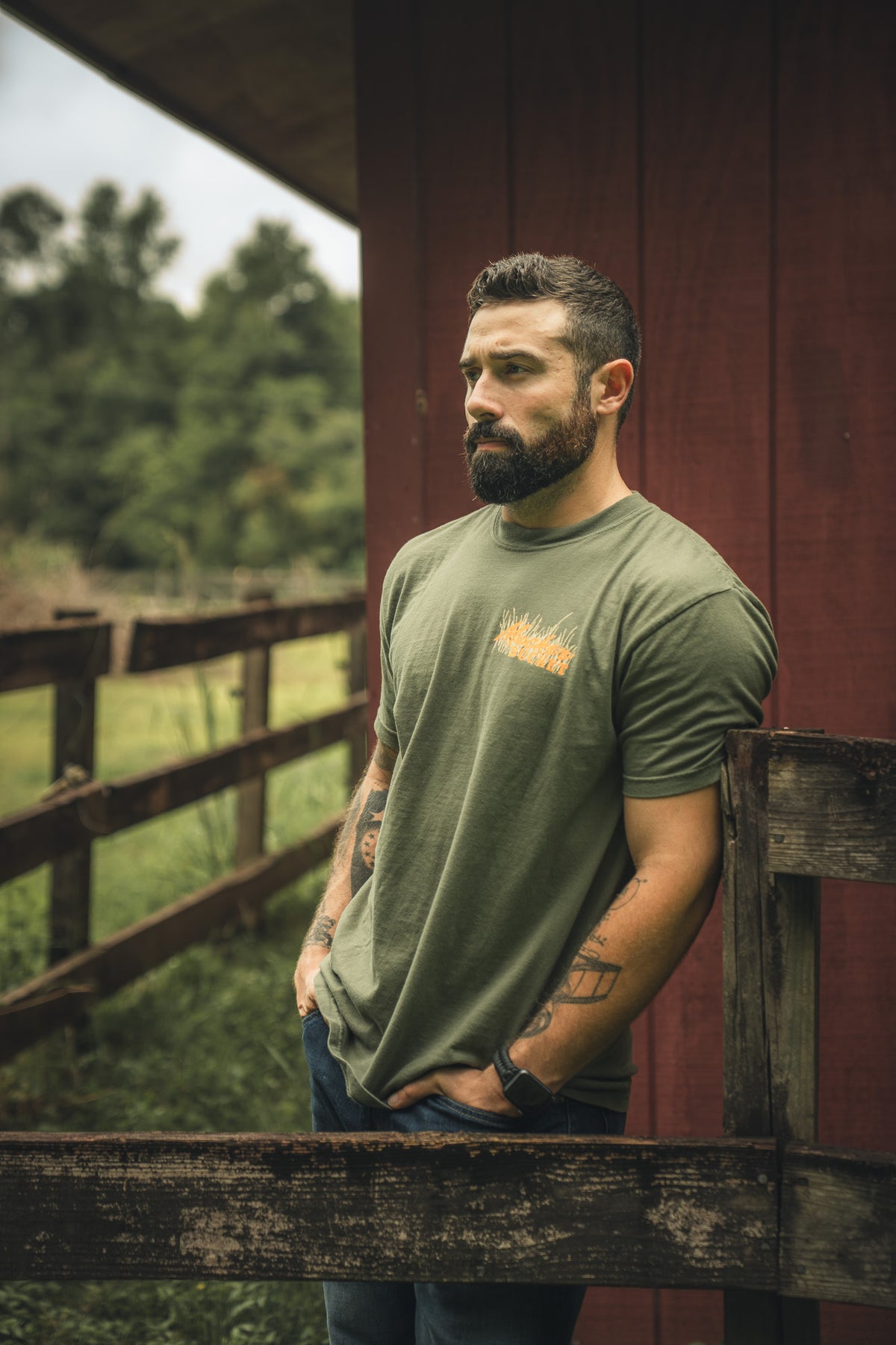 A bearded, tattooed man stands by a wooden fence outdoors, wearing Mucker Bucket’s Ghost Gobbler Premium Tee in olive green and jeans, with trees and a red barn in the background.