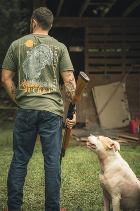 A man wears the Mucker Bucket Ghost Gobbler Premium Tee, standing on grass with his back to the camera and holding a shotgun. A light-colored dog sits nearby looking up; a wooden shed is seen in the background.