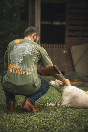 A man with a prosthetic arm kneels on grass, petting a white and tan dog. He wears jeans and a Mucker Bucket Ghost Gobbler Premium Tee in garment-dyed green, featuring a graphic design on the back. A wooden structure and greenery are behind him.