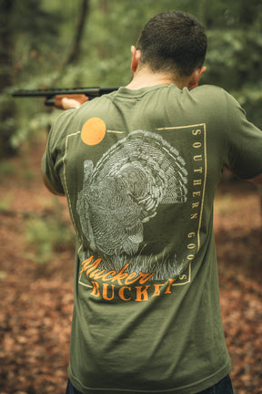 A man stands outdoors aiming a shotgun, wearing the Ghost Gobbler Premium Tee by Mucker Bucket, featuring a green color, turkey graphic, orange sun, and branded text on the back. Trees and fallen leaves are seen in the background.