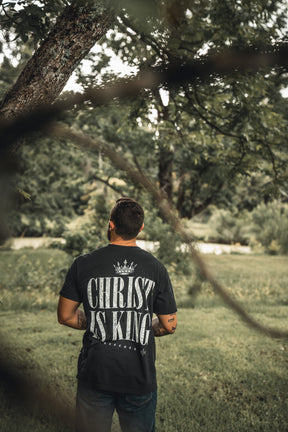 A man stands on grass near a tree, wearing the 13th brands Christ is King black T-shirt with bold white lettering on the back, set against a green, leafy background.