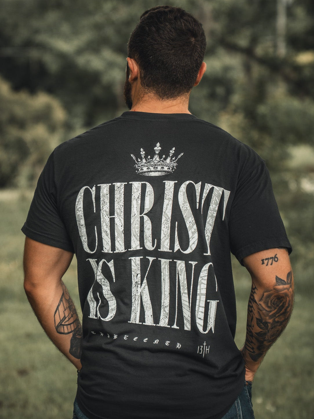 A man stands outdoors, back to the camera, wearing the 13th Christ is King black t-shirt with bold white crowned lettering. His tattooed arms are visible amidst green surroundings.