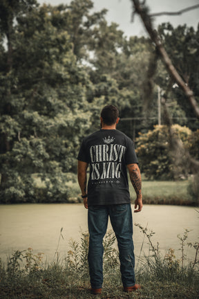A man stands by a serene pond, wearing the 13th brand Christ is King black T-shirt with large white lettering, showcasing his faith as he faces away amid lush greenery.