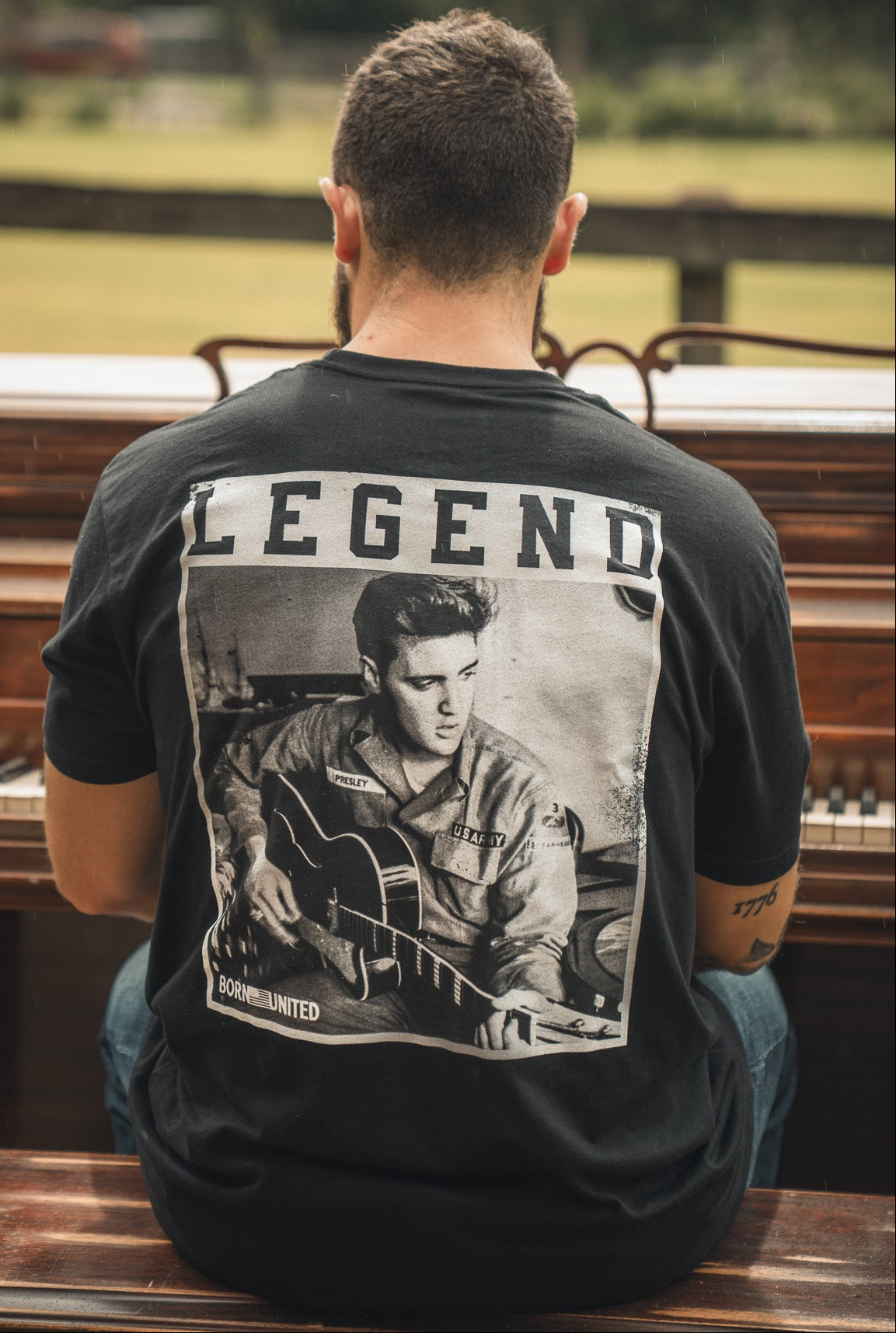 A man sits outdoors at a piano, seen from behind, wearing the Born United Private Label The King Legend Tee—a black shirt featuring a black-and-white image of Elvis Presley with LEGEND above, honoring the rock and roll icon.