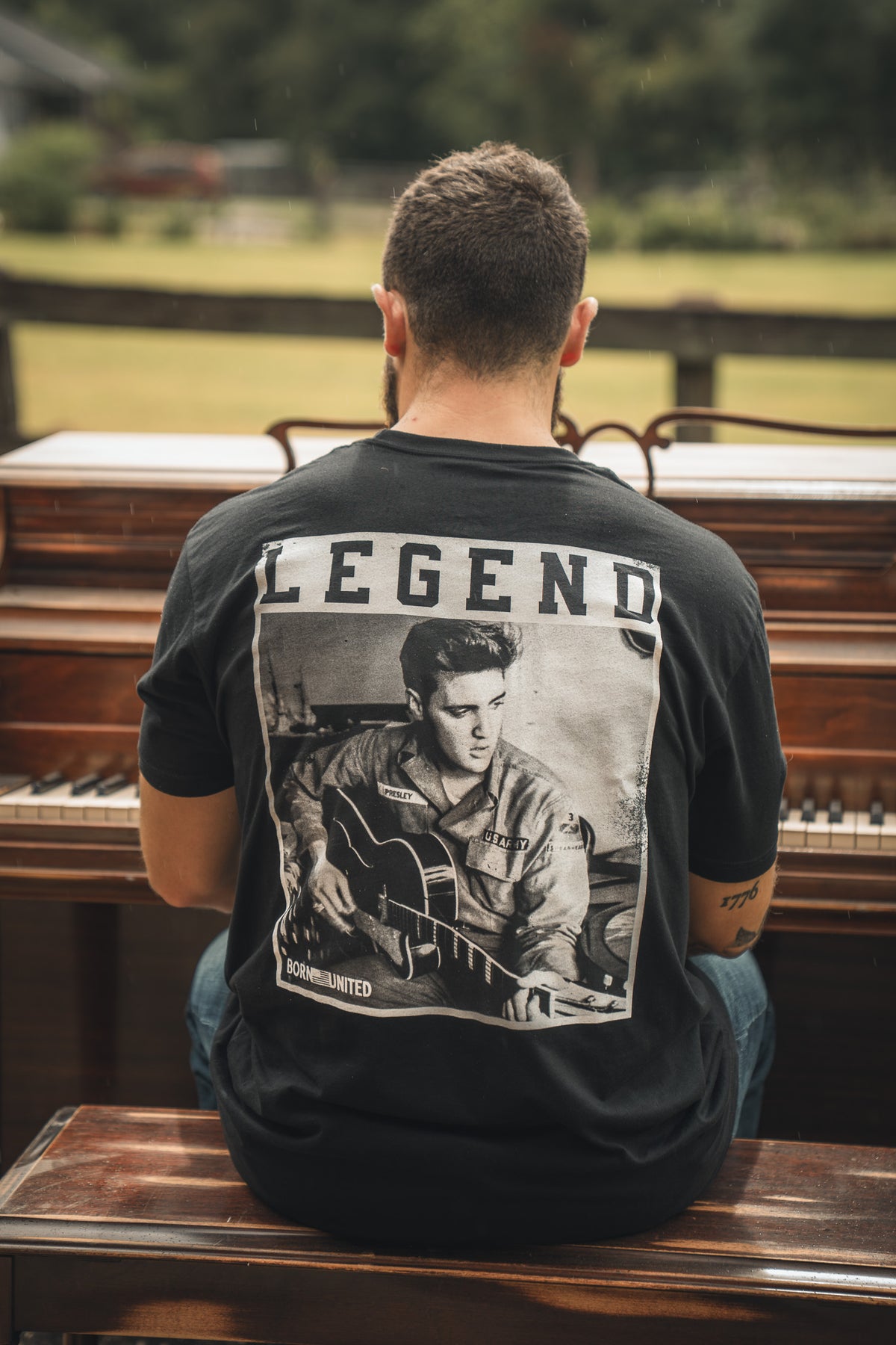 A man sits outdoors at a piano, seen from behind, wearing the Born United Private Label The King Legend Tee—a black shirt featuring a black-and-white image of Elvis Presley with LEGEND above, honoring the rock and roll icon.