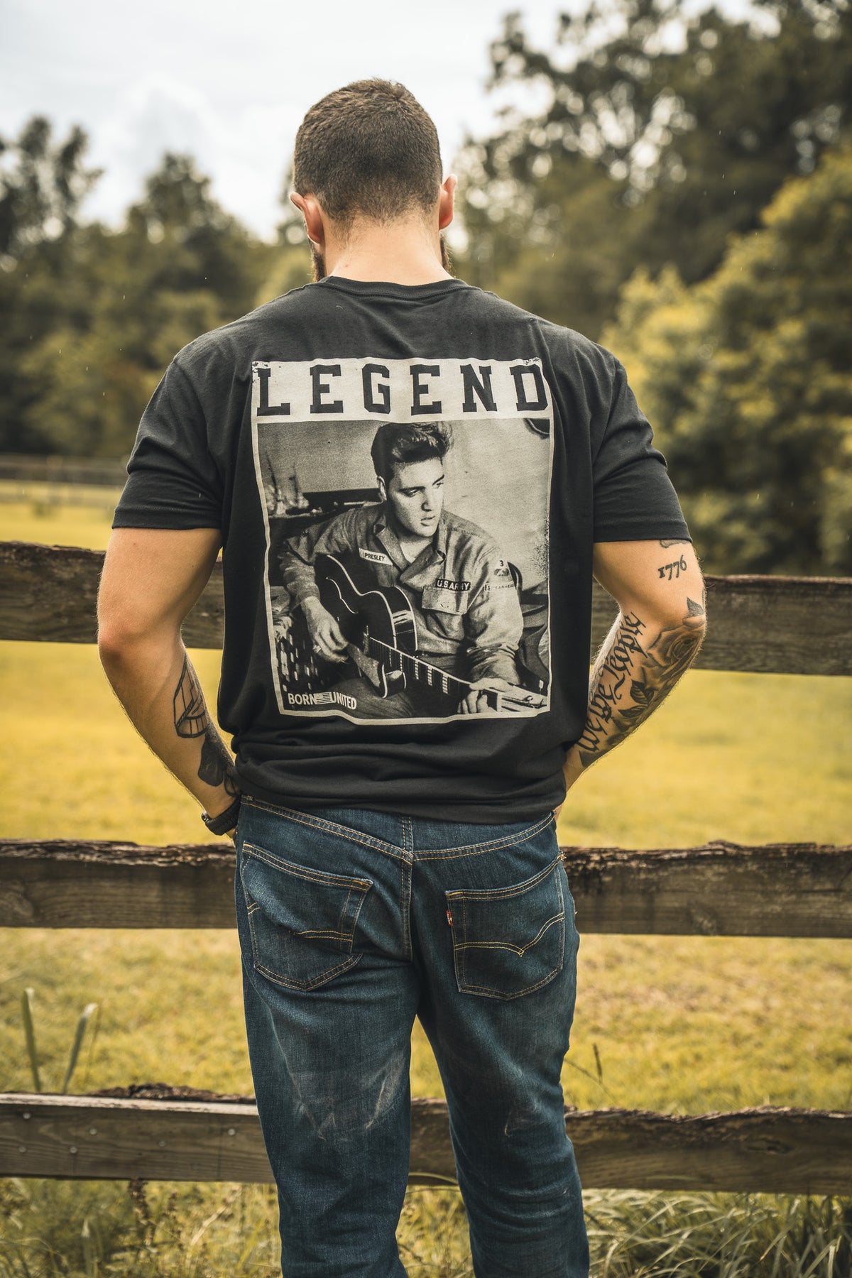A man stands by a wooden fence, facing away, wearing The King Legend Tee by Born United Private Label, featuring a rock musician photo with LEGEND above. Trees and grassy fields spread out in the background.