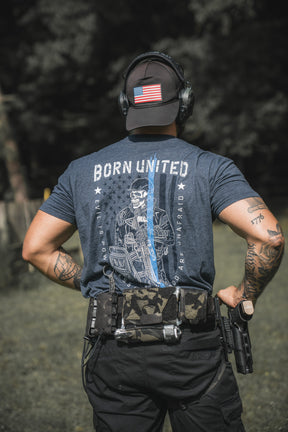 A person stands outdoors, hands on hips, wearing The Good Are Unafraid shirt by Born United Private Label with a police support graphic, plus a U.S. flag cap, headphones, and a utility belt holding gear and a firearm.