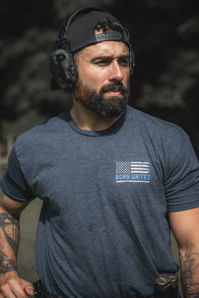A bearded man in a backward cap and headphones wears a blue The Good Are Unafraid T-shirt from Born United Private Label outdoors, showing tattoos and a holstered gun at his waist, standing strong with a serious expression.