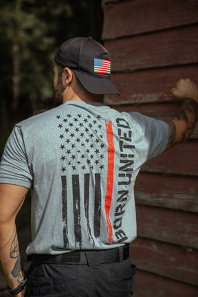 A man in a backward cap with a U.S. flag patch stands by a wooden wall, wearing the Born United Private Label Red Line Flag Tee featuring a distressed U.S. flag with a red stripe and BRAVERY printed beside it.