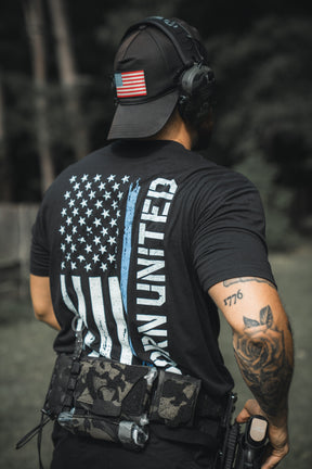 A person outdoors wears a black cap and the Born United Private Label Blue Line Flag Tee, featuring a blue line American flag and Born United text. They carry tactical gear, have a holstered weapon, and display left arm tattoos, showing Back the Blue support.