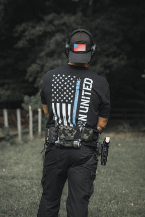 Outdoors, a person wears black tactical gear with a Born United Private Label Blue Line Flag Tee featuring a blue-striped American flag and “UNITED” text, plus an American flag patch cap, ear protection, and a holstered handgun.