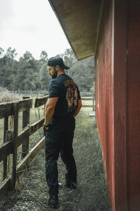 A man in the Brotherhood Forged In Flames black t-shirt by Born United Private Label, black pants, and a backward cap stands by a wooden fence near a red building, looking down with trees behind him.