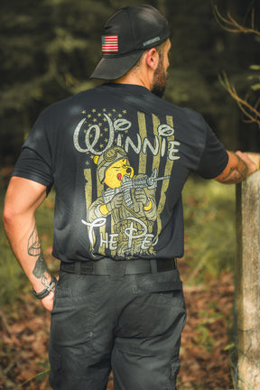 A man in a black cap and Born United Private Label’s Winnie the Pew T-shirt, which has an American flag on the sleeve and a soldier-clad bear on the back, stands outdoors blending patriotism with playful style.