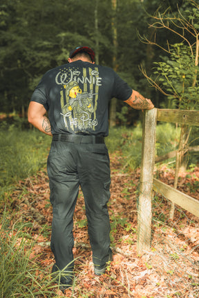 A person stands on a sunlit forest path, wearing the black Winnie the Pew t-shirt by Born United Private Label, hand resting on a wooden fence, capturing the storybook bears adventurous spirit.