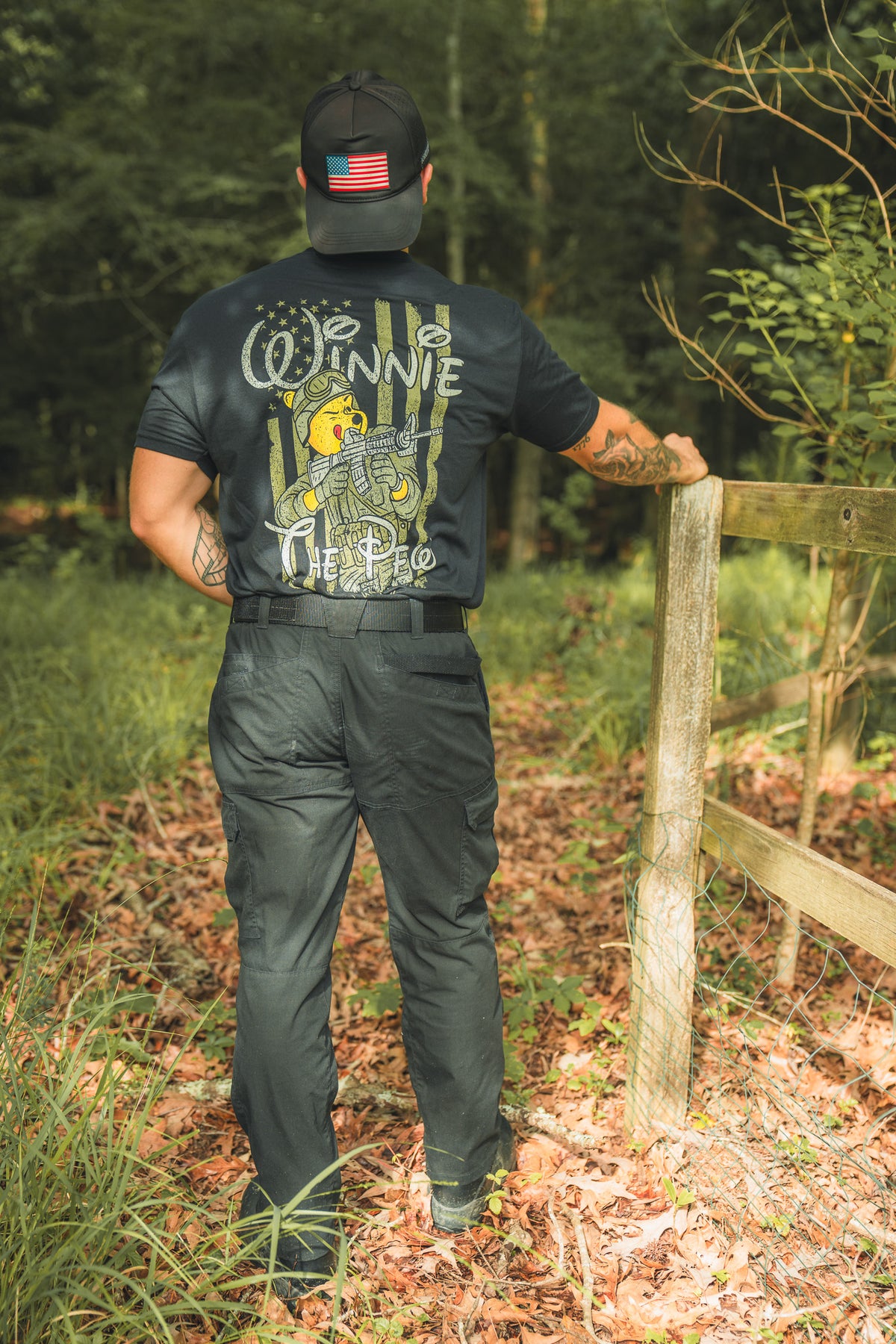 Standing outdoors by a wooden fence, this patriot wears a black Winnie the Pew shirt from Born United Private Label with a storybook bear graphic and a cap featuring an American flag patch, surrounded by trees and fallen leaves.