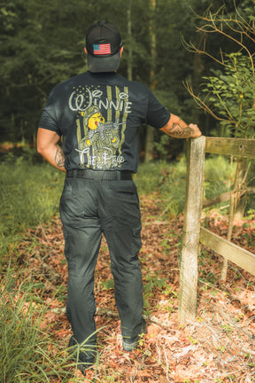 Standing outdoors by a wooden fence, this patriot wears a black Winnie the Pew shirt from Born United Private Label with a storybook bear graphic and a cap featuring an American flag patch, surrounded by trees and fallen leaves.