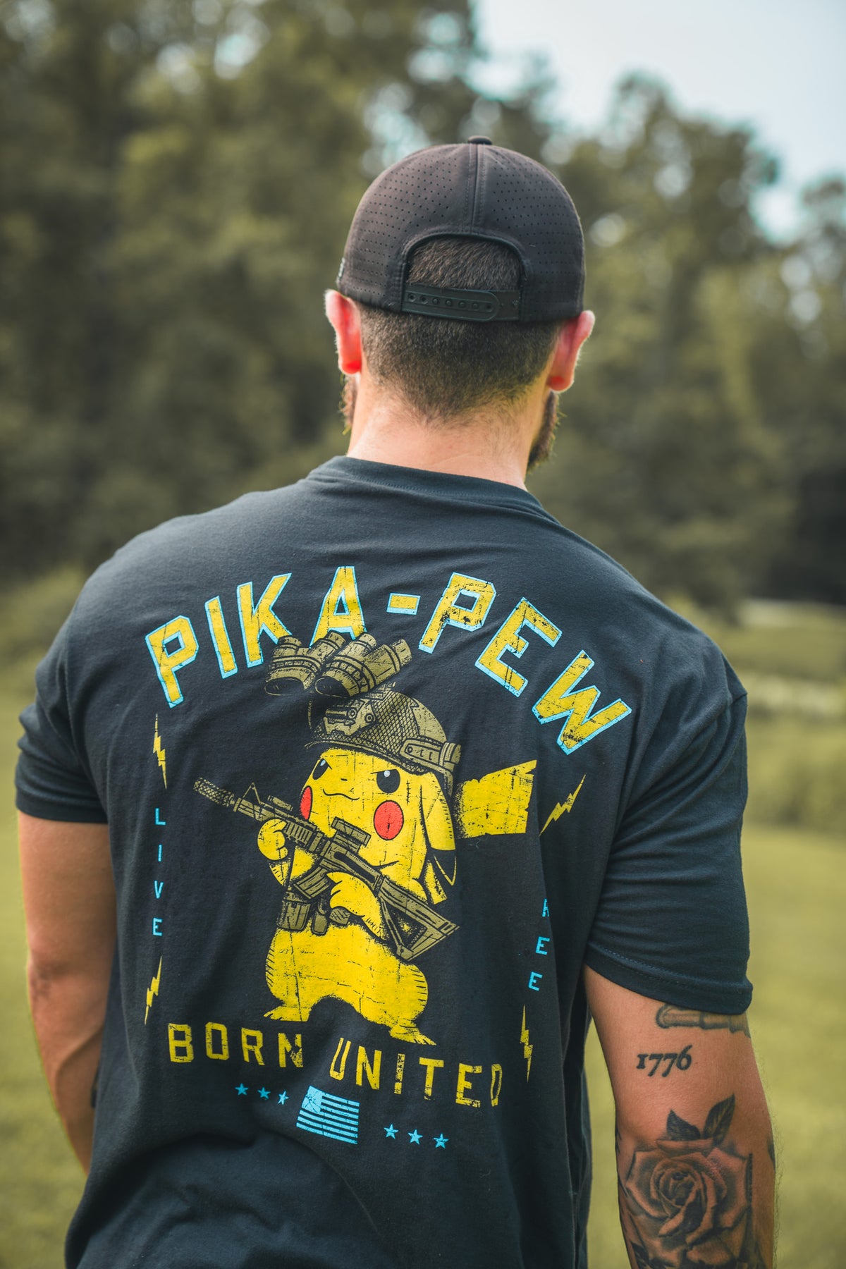 A man wears a black hat and a Born United Private Label Pika-Pew T-shirt with Pikachu holding a gun. He stands outdoors with trees behind him—a unique collectible shirt for Pokémon fans.