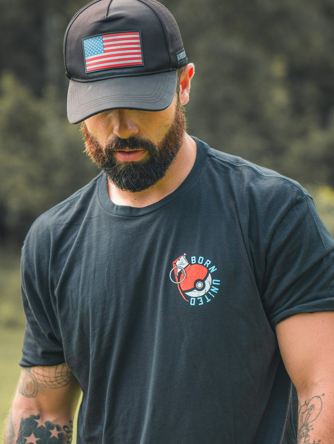 A bearded man in a black cap with an American flag patch and a Born United Private Label “Pika-Pew” tee stands outdoors, tattooed arms visible, looking down as if searching for collectibles. The background shows blurred greenery.