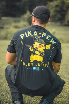 A man kneels on grass, facing away, wearing a black Born United Private Label Pika-Pew T-shirt with a Pikachu holding a rifle and bold PIKA-PEW, BORN UNITED, and LIVE FREE text—perfect for Pokémon collectors.