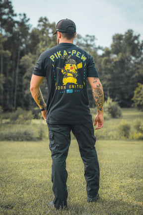 A man stands on grass, facing away, wearing a black cap, pants, and a Born United Private Label Pika-Pew T-shirt with Pikachu holding guns. Trees and greenery are in the background.