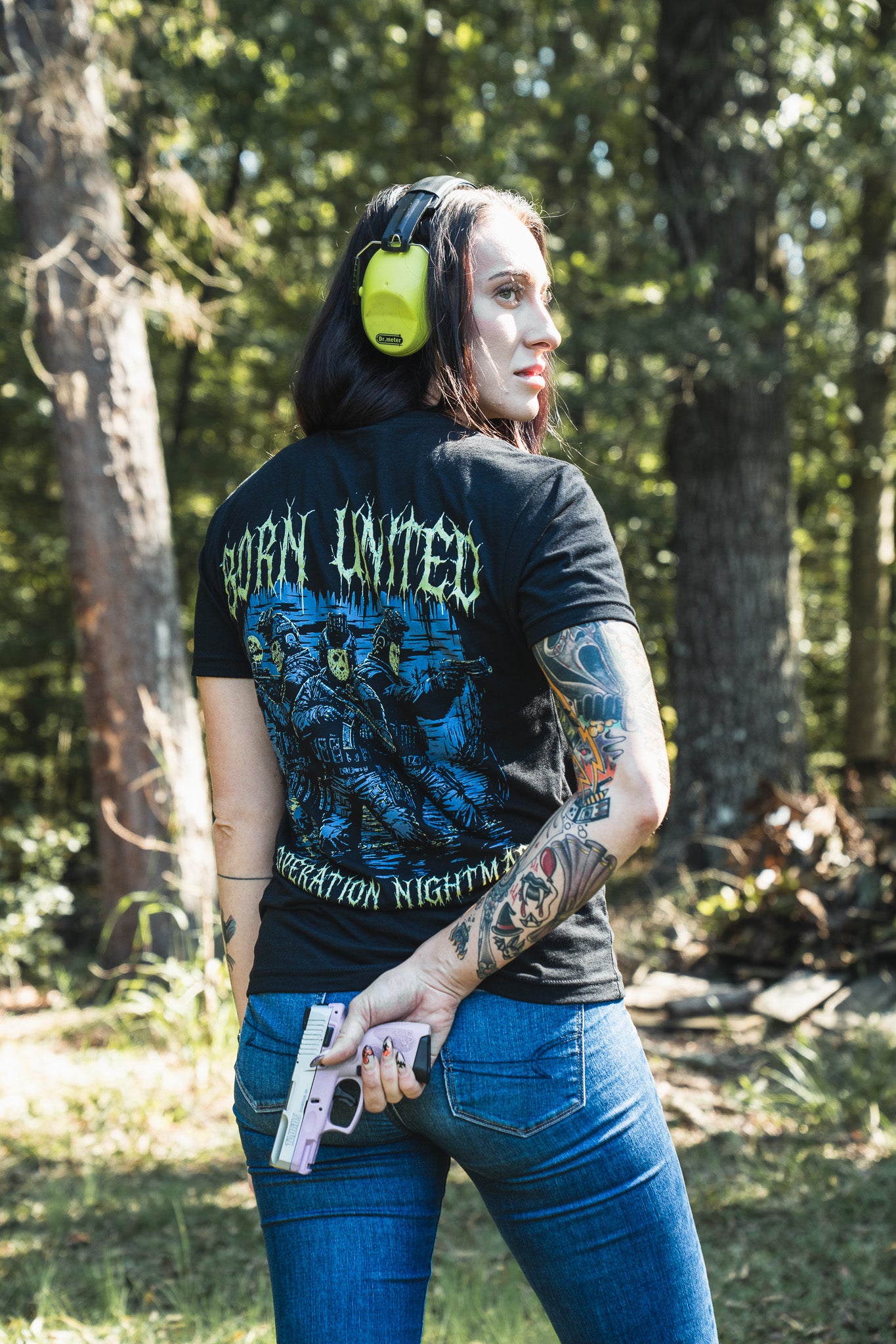 A person in blue jeans and ear protection wears a black Operation Nightmare Tee - Glow in the Dark with a soldiers graphic on the back, holding a handgun in a wooded area.
